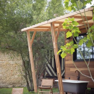 Cabane en bois avec baignoire extérieure, entourée de verdure et de chaises en bois.
