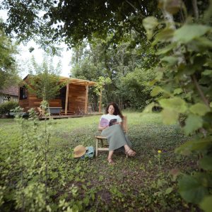Cabane en bois entourée de verdure, femme lisant paisiblement à lextérieur.