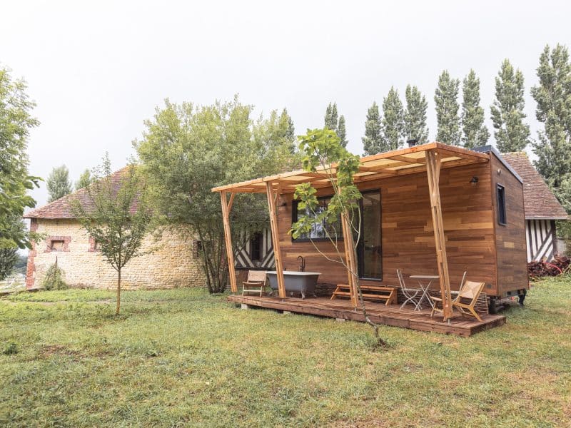 Cabane en bois avec terrasse, entourée darbres et dun jardin verdoyant.