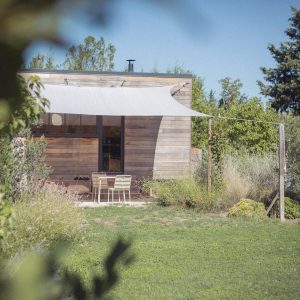 Cabane en bois avec terrasse ombragée, entourée de verdure et d'un jardin fleuri.