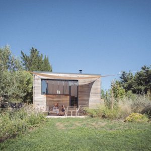 Cabane moderne en bois, entourée de verdure, avec une terrasse ombragée et un ciel bleu.