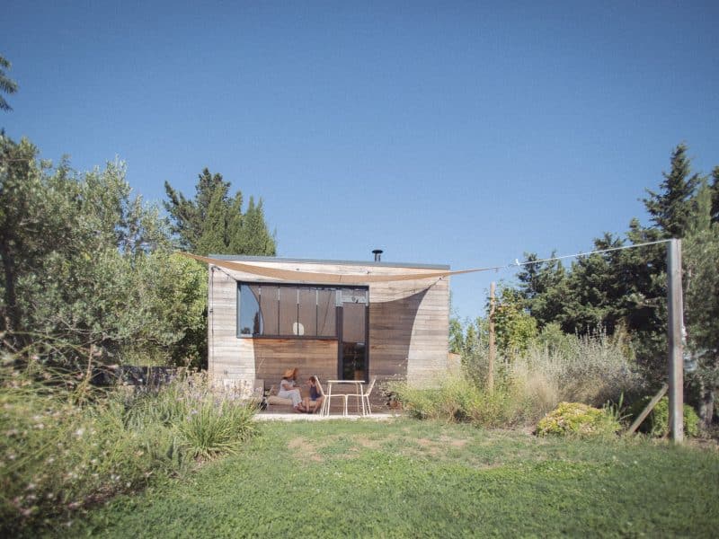 Cabane moderne en bois avec terrasse, entourée de verdure et ciel bleu.