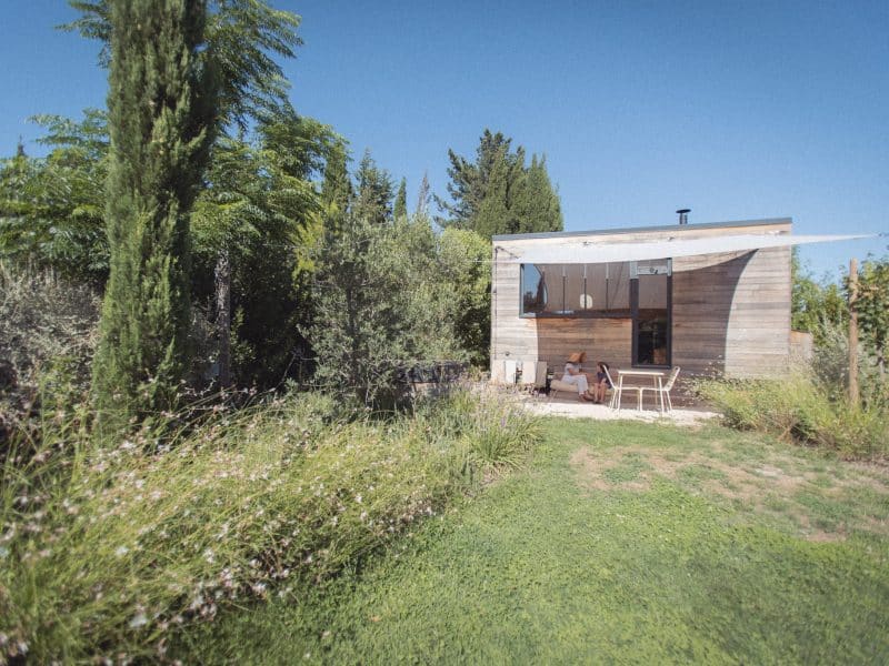 Cabane en bois moderne entourée de verdure, avec une terrasse et un ciel bleu.