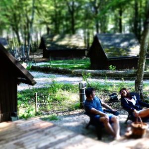 Cabane en bois dans les bois, avec des hôtes profitant d'un feu de camp. Ambiance nature apaisante.