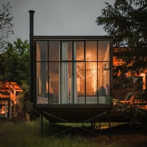 Cabane moderne en verre, illuminée par des guirlandes, entourée de verdure.