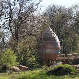 Cabane dans les arbres en forme de goutte, entourée de verdure et de rochers.