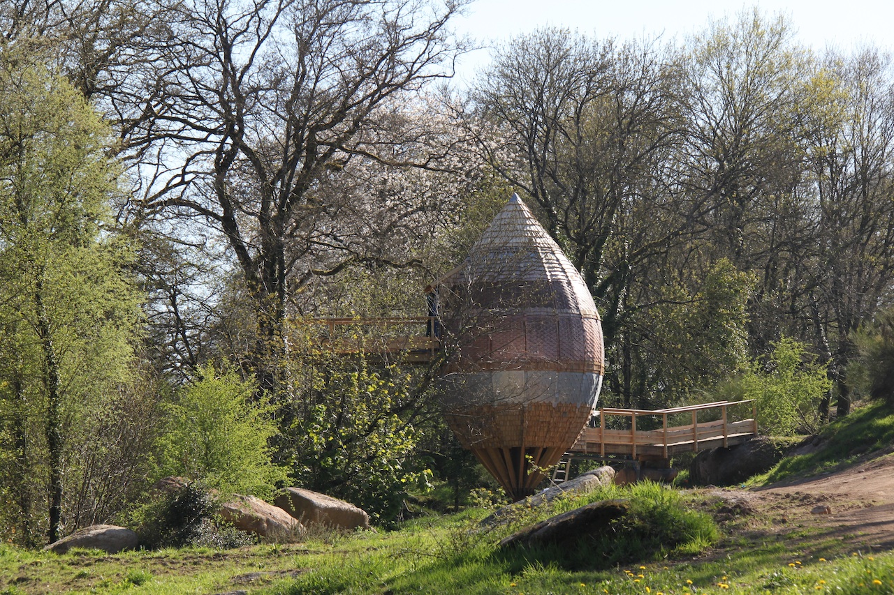 Cabane dans les arbres en forme de goutte, entourée de verdure et de rochers.