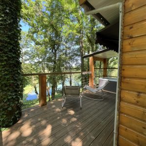Cabane en bois perchée, avec terrasse en bois et vue sur un lac entouré d'arbres verdoyants.