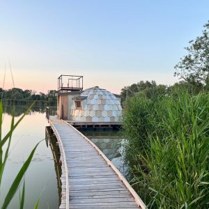 Dôme flottant sur un lac, accessible par une passerelle en bois, entouré de verdure.