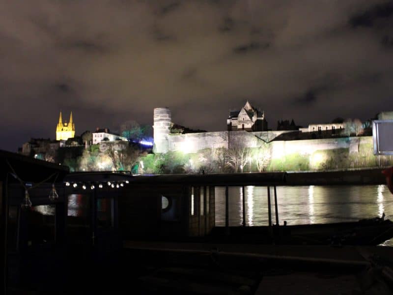 Hébergement en bord de rivière, illuminé la nuit avec un château en arrière-plan.