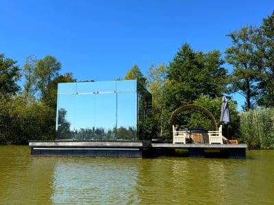 Cabane flottante en miroir, entourée d'eau et de verdure, avec un coin détente au soleil.