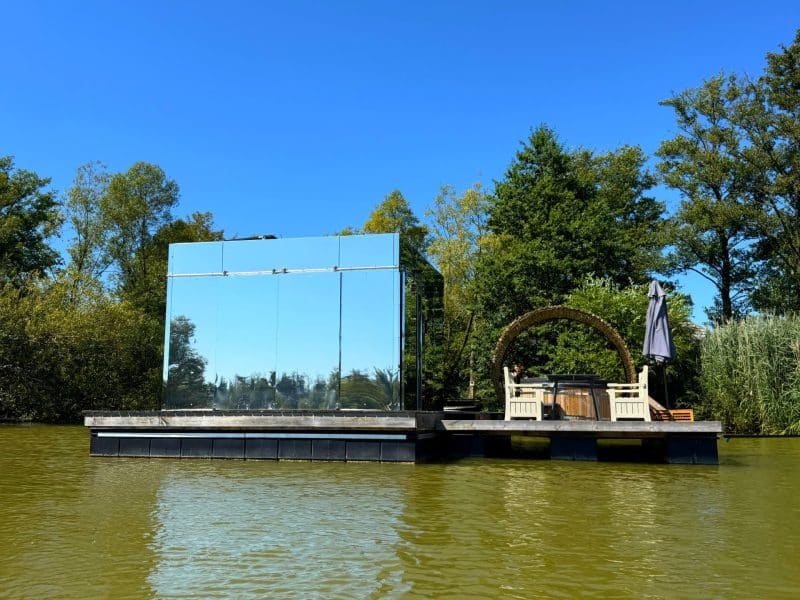 Cabane flottante en miroir, entourée d'eau et de verdure, avec un coin détente au soleil.