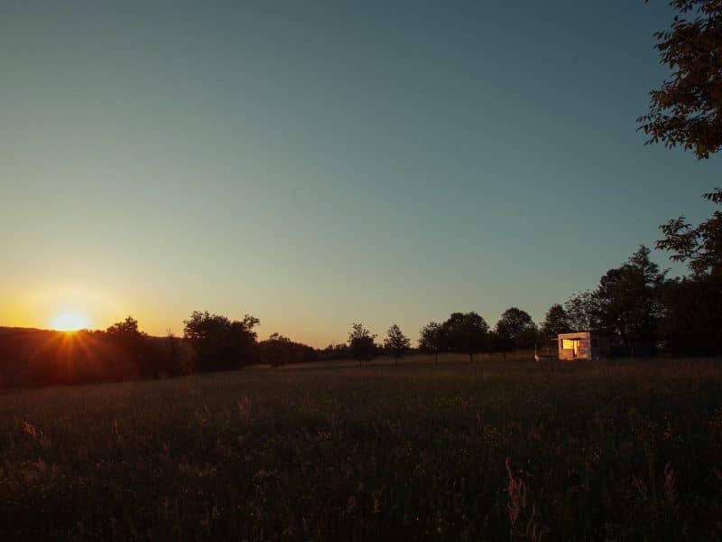 Cabane en bois au coucher de soleil, entourée de champs verdoyants et d'arbres.