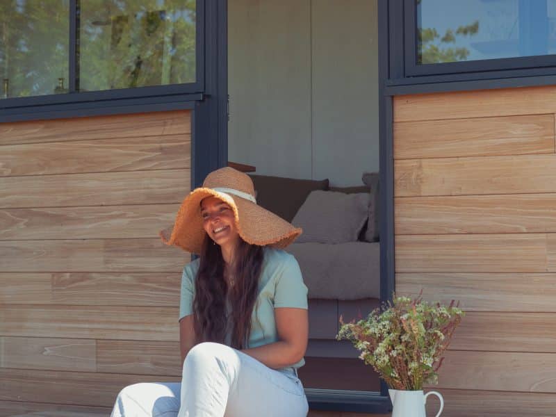 Charmante tiny house avec terrasse, une femme souriante porte un grand chapeau.