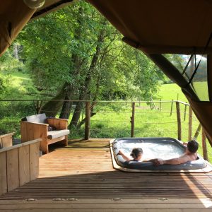 Tente de luxe avec jacuzzi sur une terrasse en bois, vue sur la nature verdoyante.