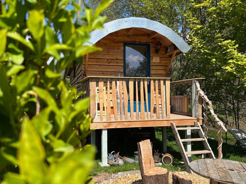 Cabane en bois perchée, avec un balcon et un coin salon en extérieur, entourée de verdure.