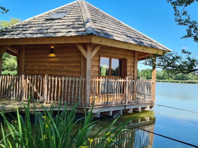 Cabane en bois sur pilotis, entourée d'eau, avec un toit en ardoise et une terrasse en bois.