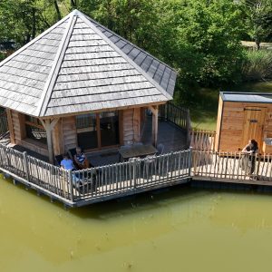 Cabane sur leau avec terrasse en bois, entourée de verdure et dun étang paisible.