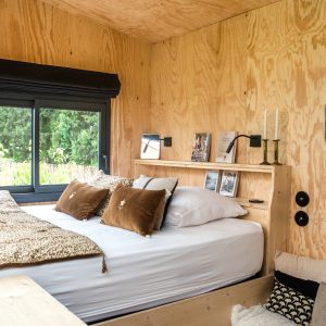 Chalet en bois minimaliste avec lit douillet, décoré de coussins et vue sur la nature.