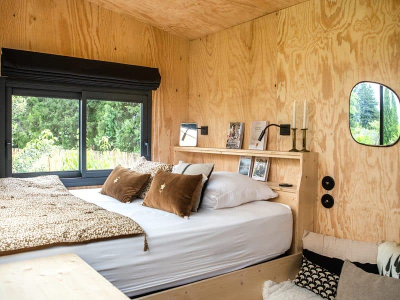 Chalet moderne en bois, lit douillet avec coussins et vue sur la nature.