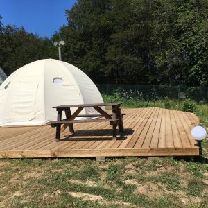 Dôme de glamping sur une terrasse en bois, entouré de verdure.