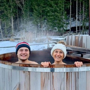 Bain nordique en bois, couple souriant dans un cadre naturel enneigé.