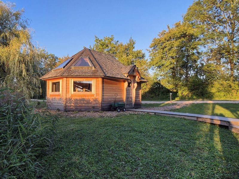 Cabane en bois ronde, entourée de verdure, avec un chemin en bois menant à l'entrée.