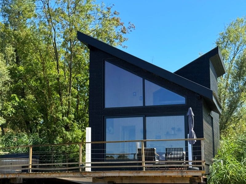Cabane moderne sur pilotis, avec grandes baies vitrées et vue sur l'eau, entourée de verdure.