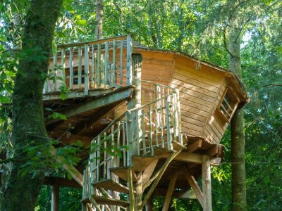 Cabane perchée dans les arbres, en bois, avec un balcon et un escalier en colimaçon.