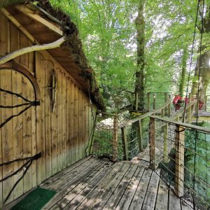 Cabane dans les arbres en bois, entourée de verdure, avec une passerelle en bois.