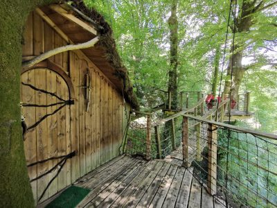 Cabane dans les arbres en bois, entourée de verdure, avec une passerelle en bois.