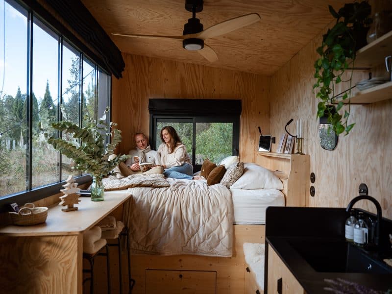Cabane moderne avec grandes fenêtres, couple détendu sur un lit douillet.