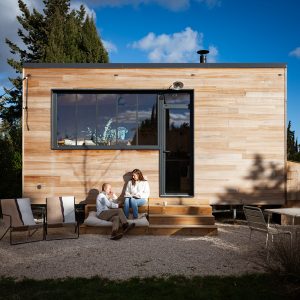 Cabane en bois moderne avec terrasse, entourée de verdure et ciel bleu. Moment de détente en couple.