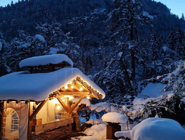 Chalet en bois sous la neige, illuminé par des guirlandes, entouré de montagnes.