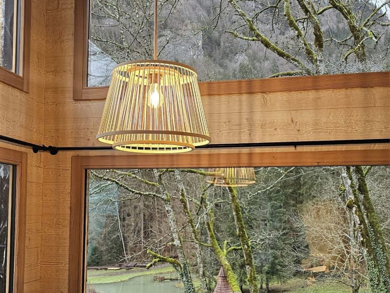 Cabane en bois avec lampes suspendues, vue sur un jardin verdoyant et un petit lac.