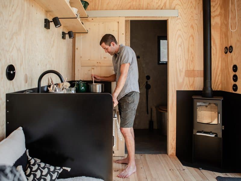 Tiny house moderne avec intérieur en bois, homme cuisinant près du poêle.