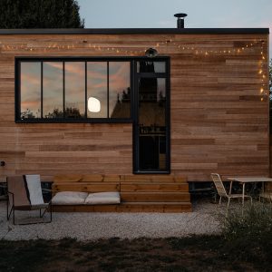 Cabane en bois moderne avec éclairage doux, terrasse accueillante et vue sur le coucher de soleil.