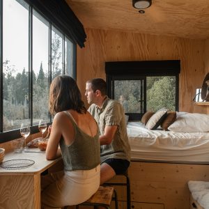 Cabane moderne en bois avec vue panoramique, couple dégustant un repas près de la fenêtre.