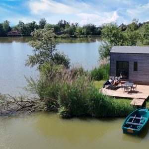 Cabane flottante au bord d'un lac, avec terrasse en bois et barque verte. Détente au soleil.