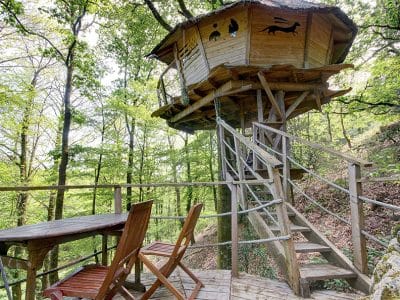 Cabane perchée dans les arbres, avec terrasse en bois et vue sur la forêt verdoyante.