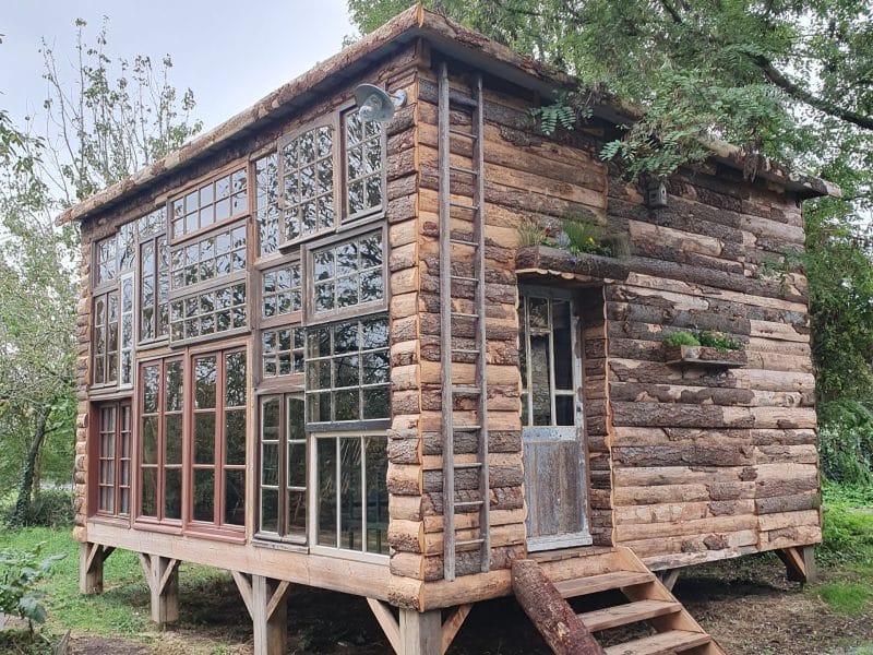 Cabane en bois avec de grandes fenêtres, entourée de verdure.