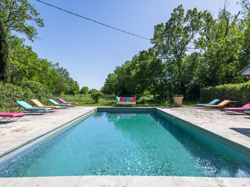 Villa avec piscine, entourée de verdure et chaises longues colorées au bord.