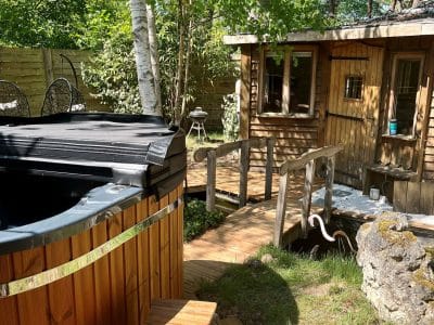 Cabane en bois avec jacuzzi, entourée de verdure et dun chemin en bois.