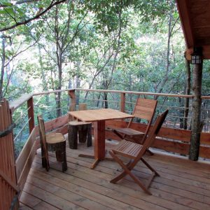Cabane dans les arbres avec terrasse en bois et chaises rustiques entourée de verdure.