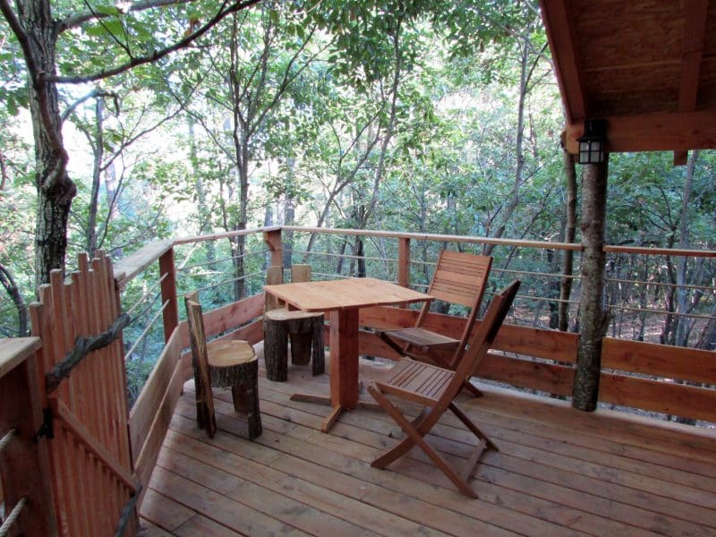 Cabane dans les arbres avec terrasse en bois et chaises rustiques entourée de verdure.