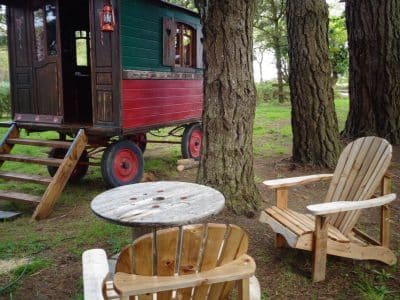 Roulotte colorée en Bretagne, entourée darbres, avec chaises en bois et table.