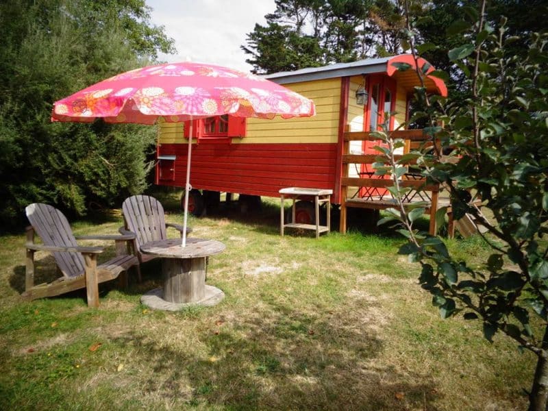 Cabane colorée en Bretagne avec parasol et chaises en bois dans un jardin verdoyant.