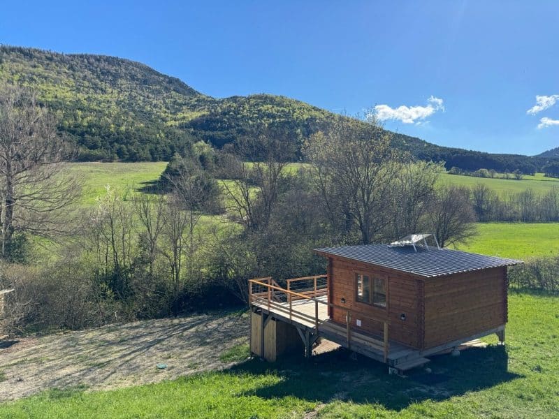 Chalet en bois avec terrasse, entouré de verdure et de montagnes ensoleillées.