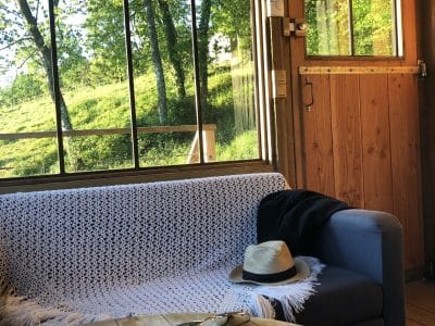 Cabane en bois avec vue sur la nature, canapé confortable et chapeau sur la table.