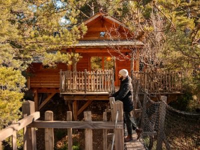 Cabane perchée en bois, entourée de verdure, accessible par un pont suspendu.